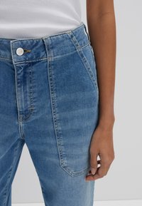 Jeans in denim blu chiaro con vita alta, caratterizzati da chiusura con bottone, tasche frontali e dettagli di cucitura a vista. Texture liscia, design casual.