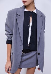 Blazer gris oversize à finition texturée, superposé à un cardigan noir à boutons, associé à une jupe grise et une ceinture noire.