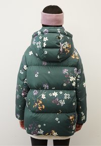 Person som bär en grön blommig pufferjacka med huva och ett rosa stickat pannband, vänd bort från kameran mot en enfärgad bakgrund.