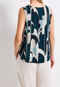 Ärmlös topp med abstrakt blå, svart och vit mönster, bärs med ljusbeiga byxor, visad bakifrån.