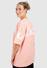 Overdimensineret T-shirt med korte ærmer i lyserød bomuld med hvid grafisk tekst på ryggen. Glat tekstur, rund halsudskæring.