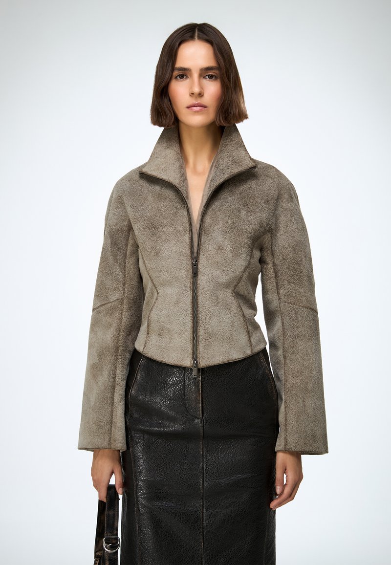 Jakna skraćenog kroja od lažnog antilopa u boji taupe s visokim ovratnikom, prednjim zatvaračem i dugim rukavima, uparena s crnom teksturiranom kožnom suknjom.