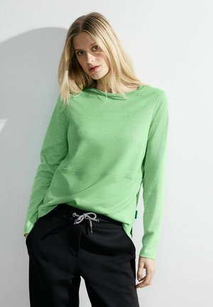 Helles grünes Langarmshirt aus strukturiertem Stoff mit rundem Ausschnitt und einer Fronttasche, kombiniert mit schwarzen Jogginghosen mit Kordelzug.