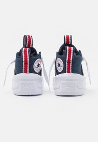 Námornické plátěné tenisky s bílými gumovými podrážkami, zdobené červenobílými pruhy a logem Converse All Star na zadní straně.