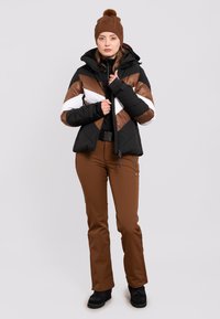Padded Jacke in Schwarz, Weiß und Braun mit Zickzackdesign, kombiniert mit braunen Hosen und einer braunen Strickmütze mit Pompon. Schwarze Stiefel.