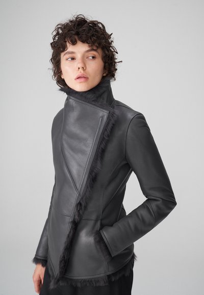 Chaqueta de cuero negra con diseño asimétrico, cuello alto y borde de piel en la parte delantera y en las mangas. Cuenta con dos bolsillos laterales.