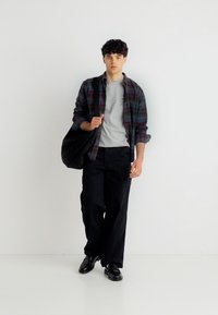 Νεαρός άνδρας περπατά προς τα εμπρός φορώντας ένα καρό πουκάμισο πάνω από γκρι μπλουζάκι, μαύρο φαρδύ παντελόνι, μαύρα παπούτσια και κρατώντας μια μεγάλη μαύρη τσάντα ώμου.