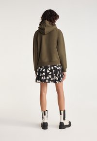 Oliwkowy hoodie z kapturem, zestawiony z czarną spódnicą w kwiaty. Model nosi masywne czarno-białe bumple; materiał wydaje się miękki i teksturowany.