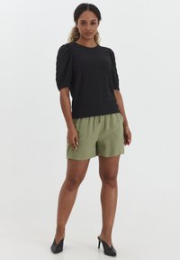 Top nero strutturato con maniche a soffietto abbinato a pantaloni corti verde oliva. Il modello indossa scarpe con tacco nere. Outfit semplice e casual.
