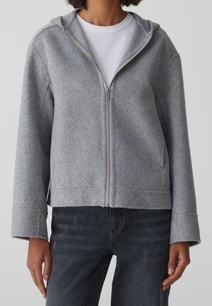 Kvinde iført en lys grå uld-hoodie med lynlås over en hvid skjorte og mørke denimjeans, stående med armene afslappet ned langs siderne.