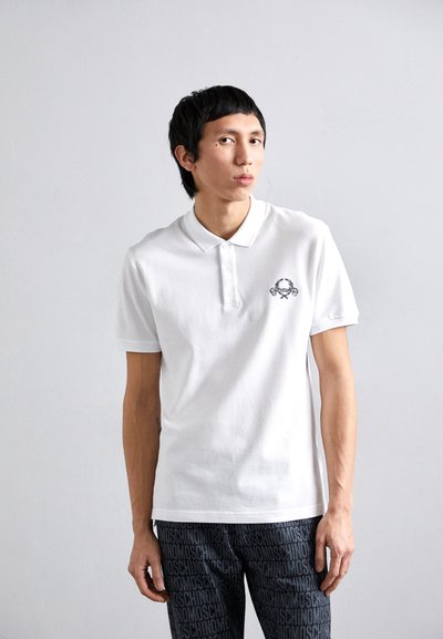 Chemise polo blanche avec patte de boutonnage à trois boutons et logo brodé sur la poitrine gauche. Manches courtes et coupe classique.