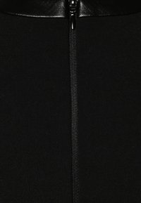 Vêtement noir ajusté avec une fermeture éclair centrale. Le matériau semble lisse avec un léger éclat au niveau de l'encolure. Tissu texturé sur les côtés.