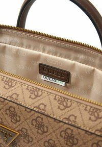 Bolso marrón con estampado y cierre de cremallera. El interior cuenta con un forro beige suave, una etiqueta de cuero con la inscripción "GUESS" y "GUESS ECO".