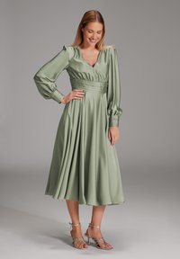 Robe midi en satin vert clair avec des manches longues, décolleté en v, taille froncée et jupe fluide. Associée à des talons argentés à lanières.