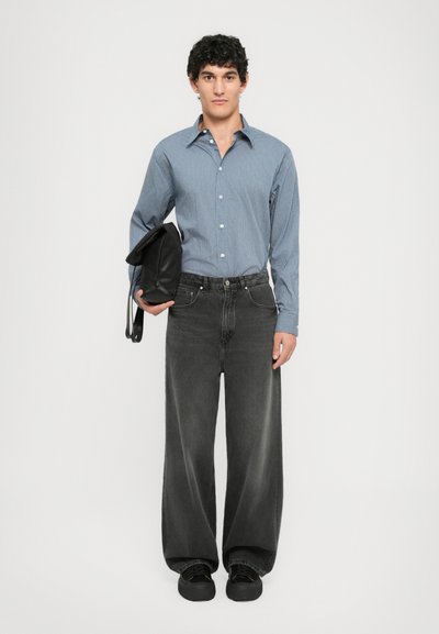 Jeune homme debout portant une chemise rayée bleue, un jean gris foncé ample, des baskets noires, et tenant un sac à dos noir sous un bras.