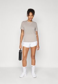 Camisa atlética de manga corta color beige con un pequeño logo, combinada con pantalones cortos blancos, botella de agua negra y zapatillas blancas con detalles en negro.