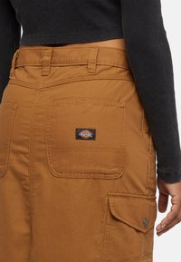 I pantaloni in tela marrone presentano una tasca posteriore con un'etichetta nera del marchio Dickies, cuciture rinforzate e una trama resistente.