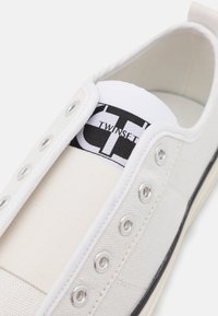 Baskets en toile blanche sans lacets, œillets argentés, logo Twinset noir et blanc sur la languette, bande noire sur la semelle.