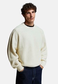 Cremfarbener Strickpullover mit strukturiertem Rippmuster, Rundhals und überschnittenen Schultern, kombiniert mit dunklen Jeans.