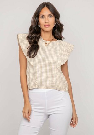 Femme aux longs cheveux bruns portant un haut sans manches en vichy beige avec des volants aux épaules et un pantalon blanc ajusté, sur fond uni.