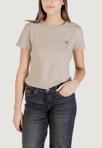 T-shirt in cotone beige chiaro con maniche corte e collo rotondo. Presenta un piccolo logo triangolare sul lato sinistro del petto. Indossato con jeans scuri.