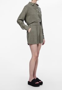 Olivově zelené šaty košilového střihu s dlouhými rukávy, zapínáním na knoflíky, elastickým pasem a kapsami. Styrováno s černými platformovými sandály. Hladká textura.