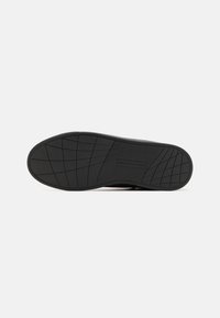 Semelle de chaussure noire avec motif texturé antidérapant et logo "Tommy Hilfiger" au centre, présentée sur un fond clair uni.