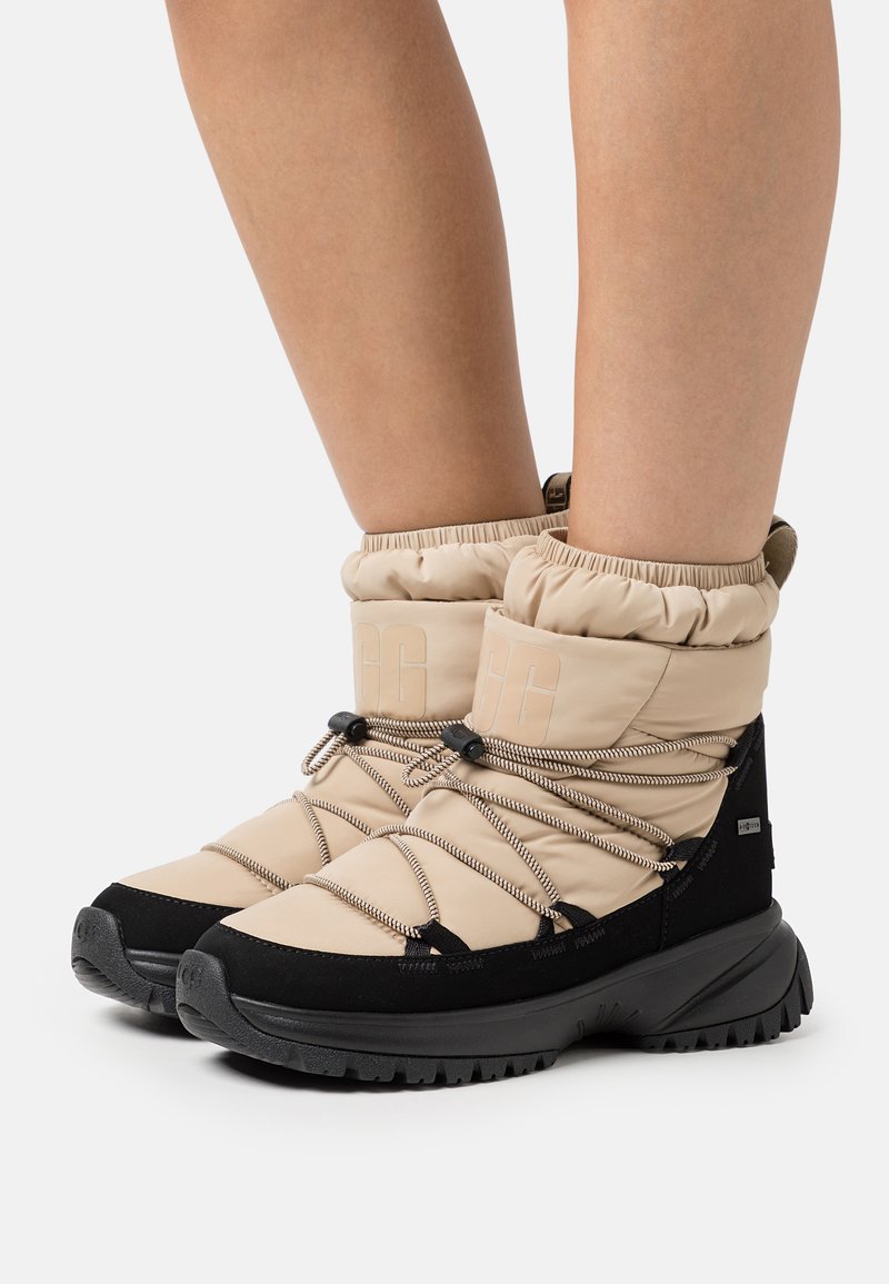 Beige gepolsterte Stiefel mit einer schwarzen Sohle, die elastische Bündchen, schwarze Akzente und graue Schnürsenkel für eine verstellbare Passform und sicheren Verschluss aufweisen.