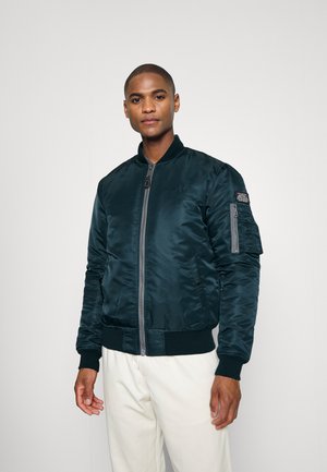 Bombér vert foncé en tissu brillant, avec une fermeture éclair sur le devant, des poignets ribbés et une poche zipée sur le bras.