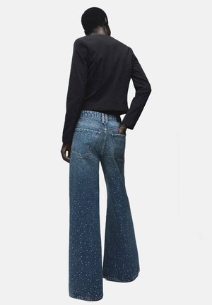 Pantalones vaqueros de tiro ancho en denim azul con un acabado desgastado y un patrón moteado, combinados con una camiseta ajustada de manga larga negra.