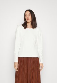 Top branco texturizado de manga longa com decote redondo e detalhes em ombreira, combinado com uma saia plissada castanha.