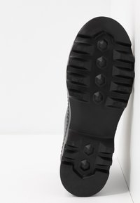 Suela de zapatilla de goma negra con un patrón de agarre texturizado, que presenta diseños hexagonales y rectangulares para tracción. Vista lateral sobre un fondo blanco.