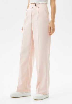 Femme portant un pantalon large taille haute rose pâle avec des plis à l'avant, un bouton noir et des baskets blanches à plateforme.