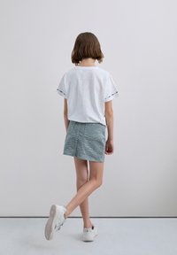 Enfant avec les cheveux bruns courts portant un t-shirt blanc et une jupe grise à motifs zébrés, s'éloignant sur un sol et un mur de couleur gris clair.
