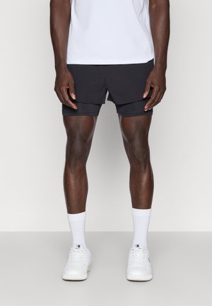 Mand iført sorte lagdelte atletikshorts, hvide crew-sokker, hvide sneakers og en hvid skjorte, stående mod en ensfarvet baggrund.