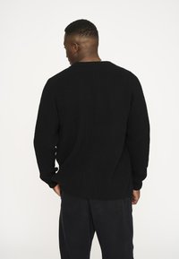 Sort ribbet sweater med lange ærmer, rund hals og sideslidser, parret med sorte, løstsiddende bukser. Tydelig tekstur i stoffet.