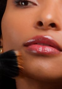 Femme aux lèvres brillantes avec un pinceau de maquillage appliquant délicatement de la poudre sur sa joue près de la bouche et du nez, gros plan.