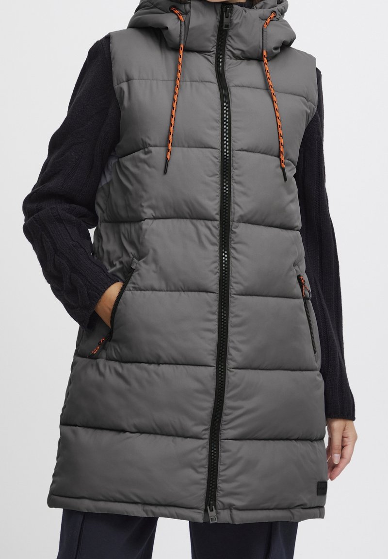 Personne portant une longue veste sans manches matelassée grise avec une fermeture éclair noire et des cordons orange sur un pull noir à manches longues.