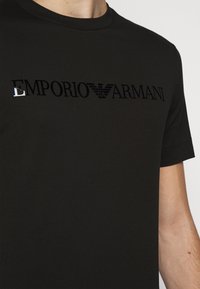 Chemise noire à manches courtes avec logo noir brillant "EMPORIO ARMANI" et emblème d'aigle sur la poitrine, portée par une personne à la peau claire.