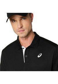 Uomo con cappellino nero e polo nera con logo bianco e pattina dei bottoni, che guarda direttamente davanti a sé su uno sfondo bianco.