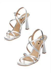 Sandalias plateadas con tacón grueso que presentan múltiples correas entrecruzadas, una punta cuadrada y un acabado metálico suave en la parte superior con una plantilla en tono marrón.