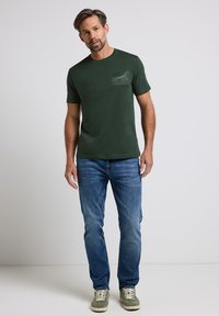 Donkergroene t-shirt met korte mouwen en een textuurzak ontwerp, gecombineerd met blauwe jeans en groene sneakers, tegen een neutrale achtergrond.