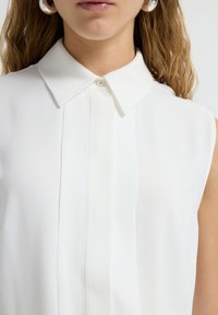 Weiße ärmellose Bluse mit klassischem Kragen, die vorne eine subtile Falte aufweist und aus einem glatten, leichten Stoff besteht.