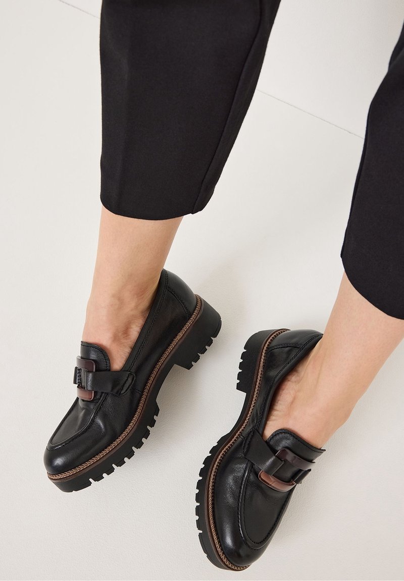 Mocassins en cuir noir avec semelles épaisses et brides décoratives à boucle, portés avec un pantalon noir raccourci sur fond clair.