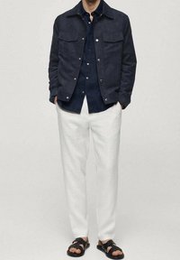 Muž ve tmavě modré košili na knoflíky a bundě, bílých kalhotách a černých sandálech stojí s rukama v kapsách na jednobarevném pozadí.