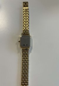 Reloj de pulsera cuadrado en tono dorado con correa de metal interconectada, parte posterior mostrando la marca Rosefield y marcas de cumplimiento sobre una superficie clara.