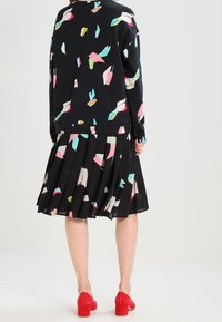 Zwarte trui met kleurrijke geometrische patronen; geplooide zwarte rok; rode schoenen met blokhak; gladde textuur; casual gelaagde stijl.