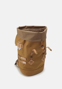 Sac à dos en toile marron avec un design à enroulement supérieur, doté d'accents en cuir, de bretelles réglables et d'une texture lisse. Style minimaliste.