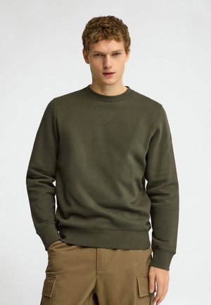 Joven con cabello corto y rizado, vestido con una sudadera verde oliva y pantalones cargo marrón claro, de pie delante de un fondo liso.