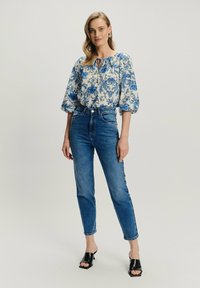 Mujer vistiendo una blusa floral azul y unos jeans de tiro alto azules, con sandalias de tacón negras, de pie contra un fondo claro y sencillo.